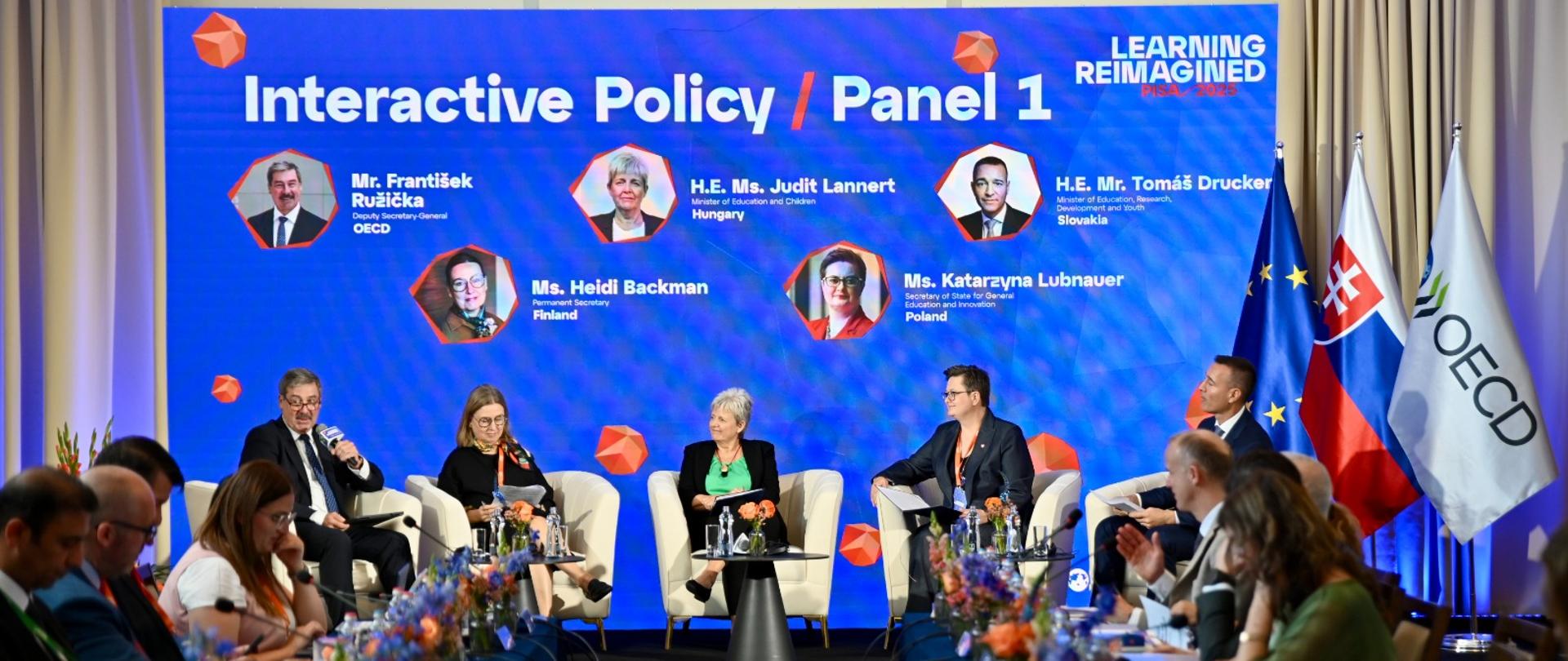 Na scenie siedzi 5 osób, w tym wiceminister Katarzyna Lubnauer, którzy uczestniczą w panelu dyskusyjnym. Przed nimi stoją stoliki z kwiatami oraz wodą. Przed sceną zasiadają uczestnicy konferencji siedzący przy dwóch stołach ustawionych prostopadle do sceny. Po prawej stronie sceny stoją flagi Unii Europejskiej, Słowacji i OECD.