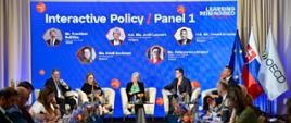Na scenie siedzi 5 osób, w tym wiceminister Katarzyna Lubnauer, którzy uczestniczą w panelu dyskusyjnym. Przed nimi stoją stoliki z kwiatami oraz wodą. Przed sceną zasiadają uczestnicy konferencji siedzący przy dwóch stołach ustawionych prostopadle do sceny. Po prawej stronie sceny stoją flagi Unii Europejskiej, Słowacji i OECD.