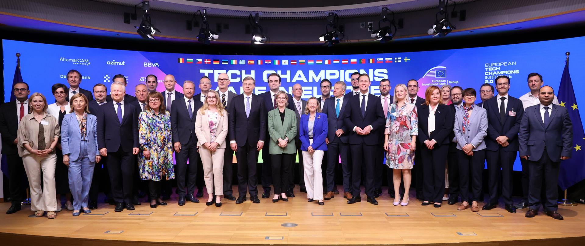  Uczestnicy wydarzenia European Tech Champions Initiative pozują do wspólnego zdjęcia na scenie przed ekranem z nazwą inicjatywy, w otoczeniu flag państw europejskich i Unii Europejskiej.