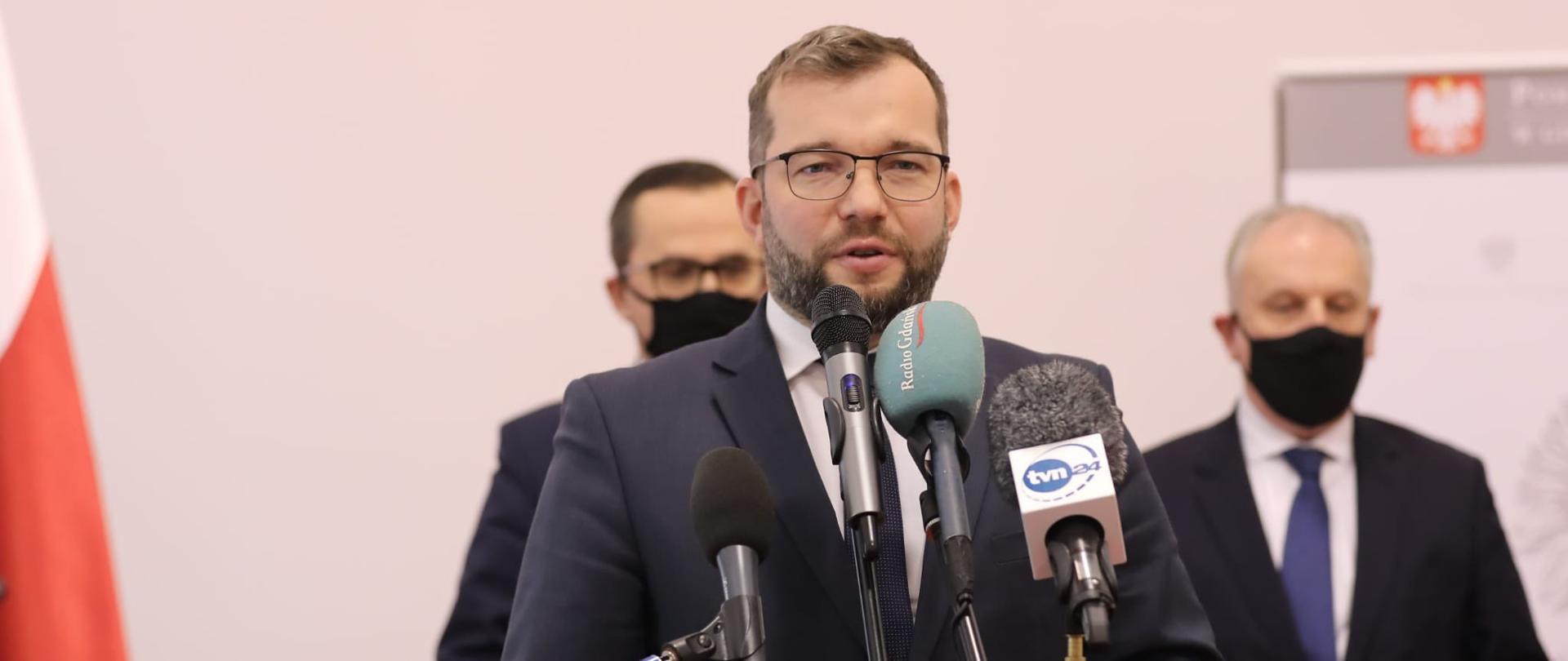 Minister Grzegorz Puda przed mikrofonami. Za nim dwóch mężczyzn. Obok flaga Polski.
