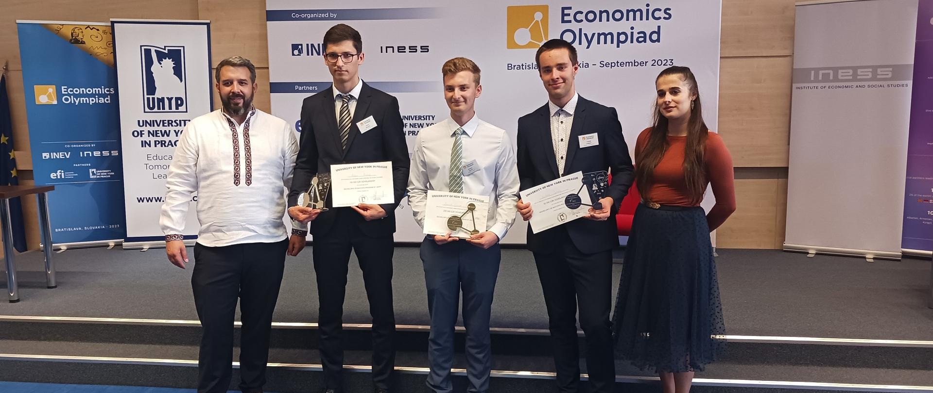 Pod ścianą na której wisi baner z napisem Economics Olympiad stoi czterech mężczyzn i kobieta, trzech w środku trzyma dyplomy.