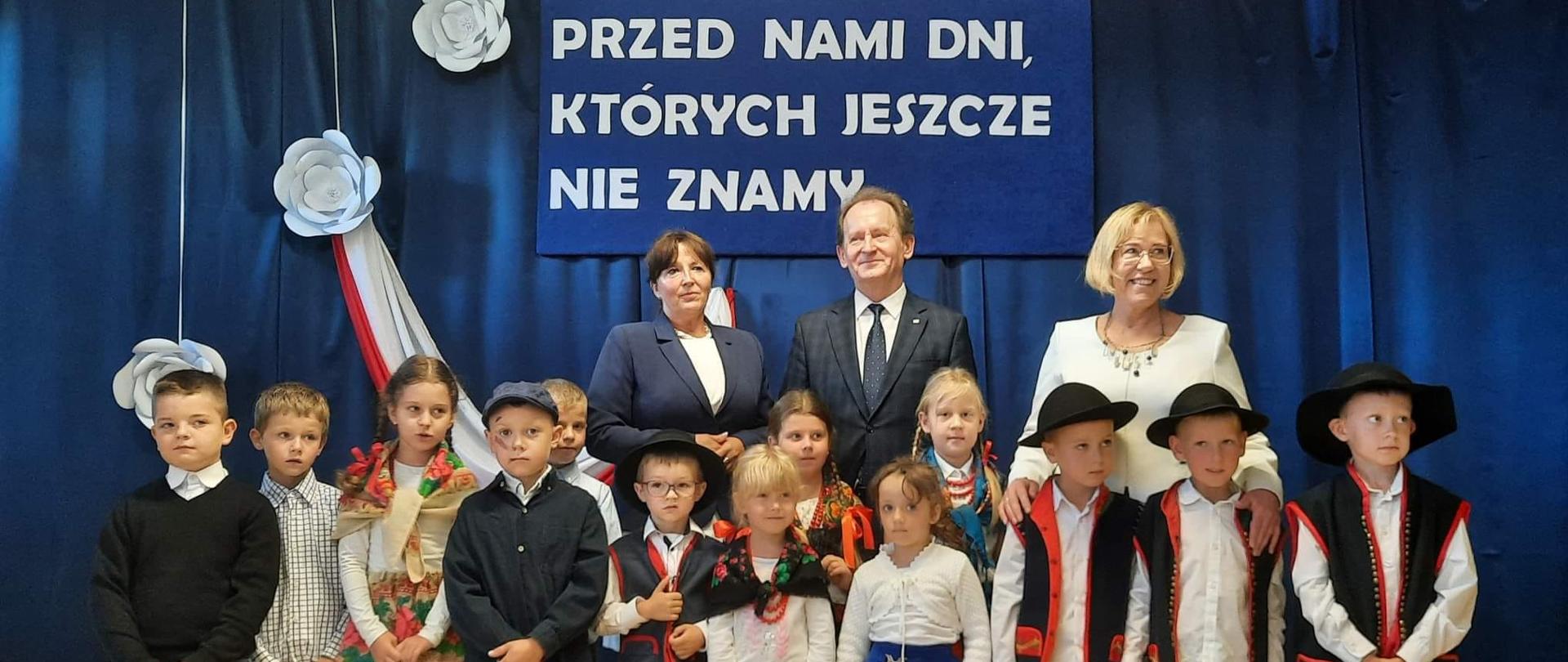 Grupa dzieci ubranych w góralskie stroje. Za nimi dyrektor szkoły, wiceminister Włodzimierz Bernacki oraz kurator Barbara Nowak. Granatowe tło i napis przed nami dni, którycj jeszcze nie znamy. 