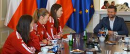Minister Sportu i Turystyki Kamil Bortniczuk siedzi przy owalnym stole wraz z polskimi szpadzistkami w składzie: Martyna Swatowska-Wenglarczyk, Ewa Trzebińska i Magdalena Pawłowska. Zawodniczki siedzą w czerwonych dresach z polskim godłem i opowiadają o niedawno zdobytym złotym medalu na mistrzostwach świata w Mediolanie. Na stole znajduje się słodki poczęstunek, woda, soki oraz kawa i filiżanki. W tle stoją flagi Polski i Unii Europejskiej, oraz wisi lustro nad kominkiem.