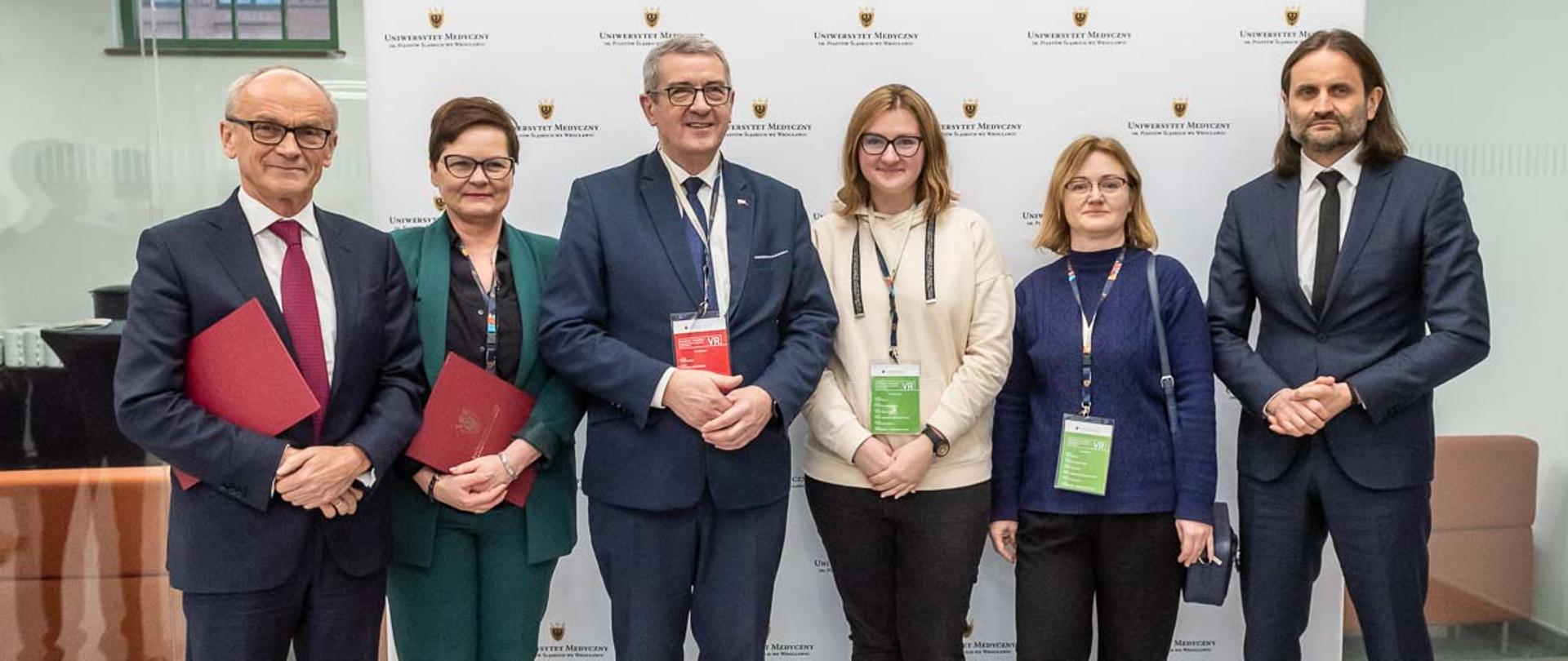 Trzy kobiety i trzech mężczyzn w garniturach stoi na tle białej ściany, dwoje trzymają czerwone teczki z godłem, wszyscy się uśmiechają.