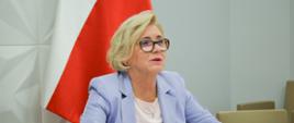 Wiceminister Machałek siedzi przy biurku, za nią przy ścianie polska flaga.