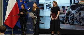 Minister funduszy i polityki regionalnej Katarzyna Pełczyńska-Nałęcz i wiceministra Monika Sikora podczas konferencji prasowej poświęconej inkubatorom na innowacje społeczne, panie minister stoją na scenie przy mikrofonach, obok minister Pełczyńskiej-Nałęcz stoi tłumaczka języka migowego, za ich plecami zdjęcie przedstawiające młodego mężczyznę na wózku inwalidzkim