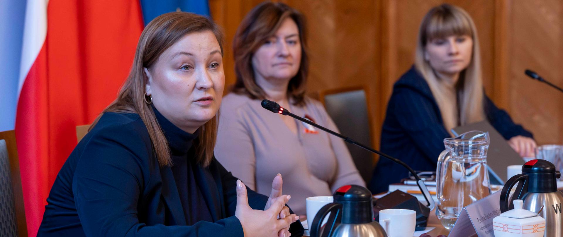 Zdjęcie przedstawia Paulinę Piechnę-Więckiewicz, podsekretarz stanu w MEN, przemawiającą przez mikrofon. Przed nią stoją filiżanki, owoce, pączki i termosy. Obok niej siedzą dwie postacie, w tym Rzeczniczka Praw Dziecka.