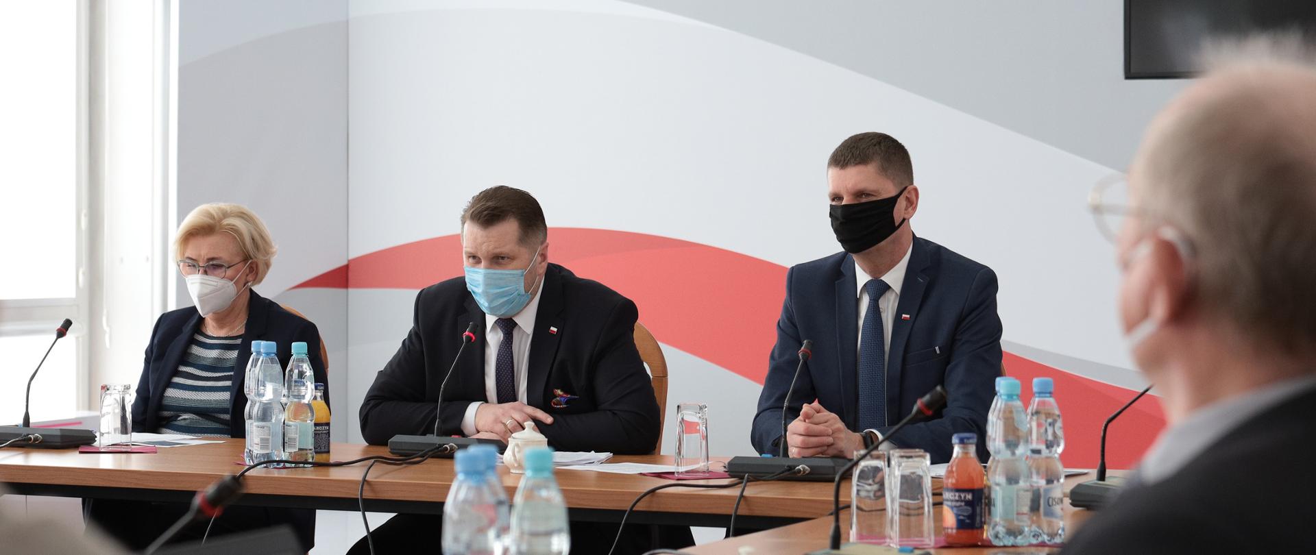 Przy stole na drugim tle siedzą wiceminister Marzena Machałek, minister Przemysław Czarnek oraz wiceminister Dariusz Piontkowski. Wszyscy mają na twarzach maseczki. W tle ściana z biało-czerwonymi barwami. Na stole butelki z wodą. Na pierwszym planie plecy osób, które siedzą prostopadle do członków kierownictwa MEiN.