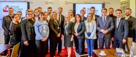 Zdjęcie zbiorowe, w sali stoi duża grupa ludzi, za nimi na niskiej ściance monitory z napisem Senat Polonii Europy, dalej żółta ściana.