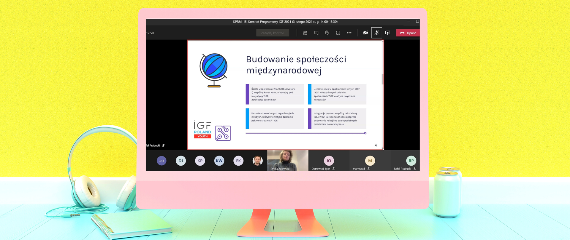 Różowy komputer na tle żółtej ściany stoi na pistacjowym biurku. Obok niego pistacjowe słuchawki, notes i puszka z napojem.