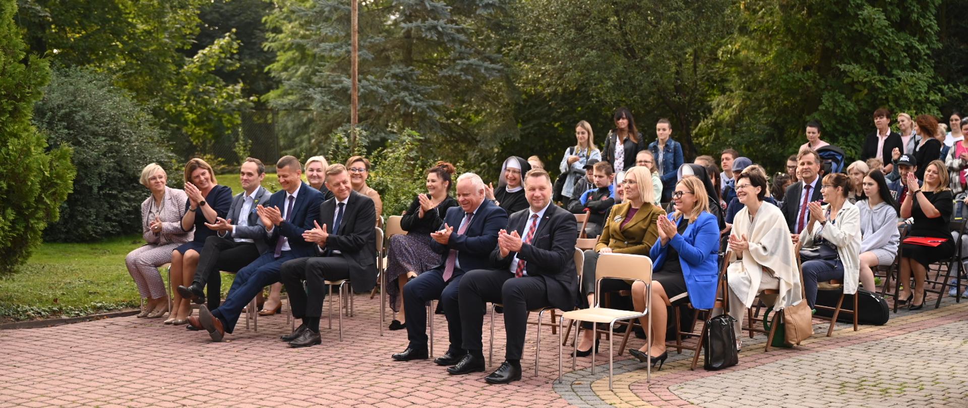 Widok z przodu pod kątem, na wyłożonym szaro-czerwoną kostką placyku na ustawionych w rzędy krzesłach siedzą ludzie, za nimi zielone drzewa.