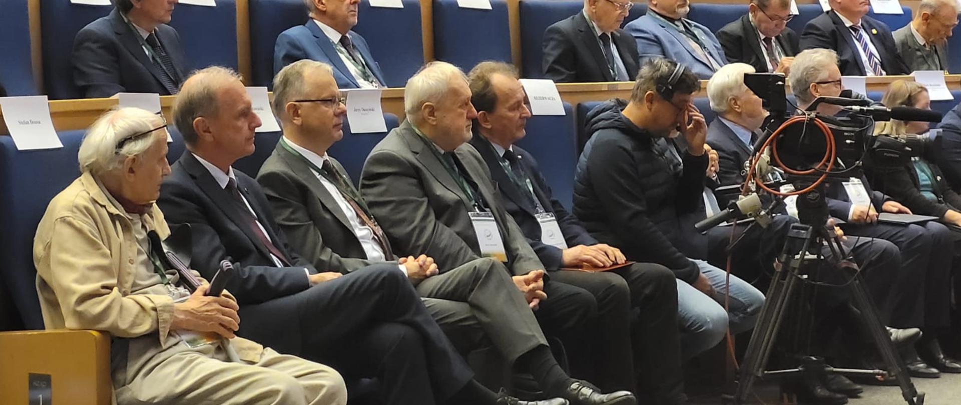 Widok z boku na dużą salę, na krzesłach siedzą ludzie.