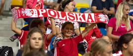 Na stadionowej trybunie siedzą dzieci, dwóch chłopców w czerwonych koszulkach trzyma rozpostarty biało-czerwony szalik z napisem Polska.
