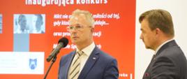 Dyrektor Adamiec stoi i mówi do mikrofonu, za nim czerwona ścianka z napisem Konferencja prasowa inaugurująca konkurs, z boku stoi mężczyzna w czarnym garniturze.