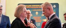 Mężczyzna w garniturze stoi naprzeciwko kobiety w czarnym ubraniu, do którego coś jej przypina.