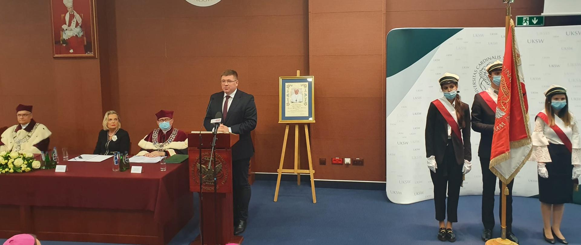Inauguracja roku akademickiego 2021/2022 na UKSW z udziałem Sekretarza Stanu w MEiN, Tomasza Rzymkowskiego. wiceminister przemawia do mikrofonu przy mównicy, z prawej strony poczet sztandarowy. 