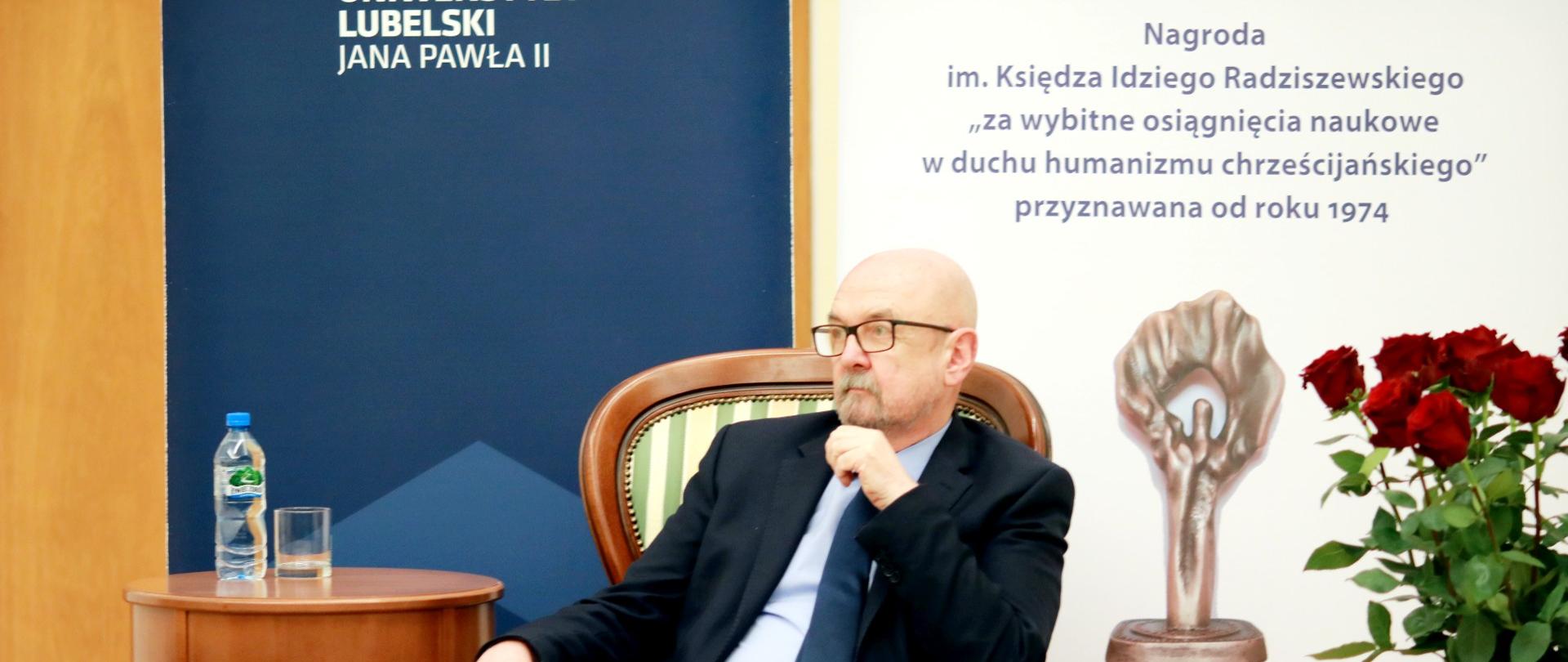 prof. legutko siedzi na ozodbnym krześle w zielone pasy, obok stolik z wodą i szklanką, po prawej stronie baner z napisem Nagroda im. Księdza Idziego Radziszewskiego "za wybitne osiągnięcia naukowe w duchu humanizmu chrześcijańskiego", obok bukiet czerwonych róż w wazonie 