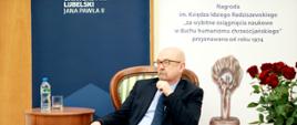 prof. legutko siedzi na ozodbnym krześle w zielone pasy, obok stolik z wodą i szklanką, po prawej stronie baner z napisem Nagroda im. Księdza Idziego Radziszewskiego "za wybitne osiągnięcia naukowe w duchu humanizmu chrześcijańskiego", obok bukiet czerwonych róż w wazonie 