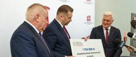 Minister Czarnek stoi pod białą ścianą, razem z mężczyzną w garniturze trzyma wielki symboliczny czek z napisem 1 734 000 zł, obok stoi inny mężczyzna w garniturze.