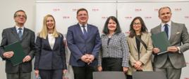 Zdjęcie zbiorowe, trzech mężczyzn i trzy kobiety stoją pod ścianą i uśmiechają się, przed nimi stół przykryty czarnym materiałem.