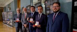 Pod szarą ścianą stoi grupa mężczyzn w garniturach, trzymają małe pudełeczka w których widać medale.