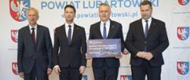 Pod ścianką z napisem Powiat Lubartowski stoi minister Czarnek i trzech mężczyzn w garniturach, jeden trzyma tabliczkę z napisem Gmina Ostrów Lubelski.