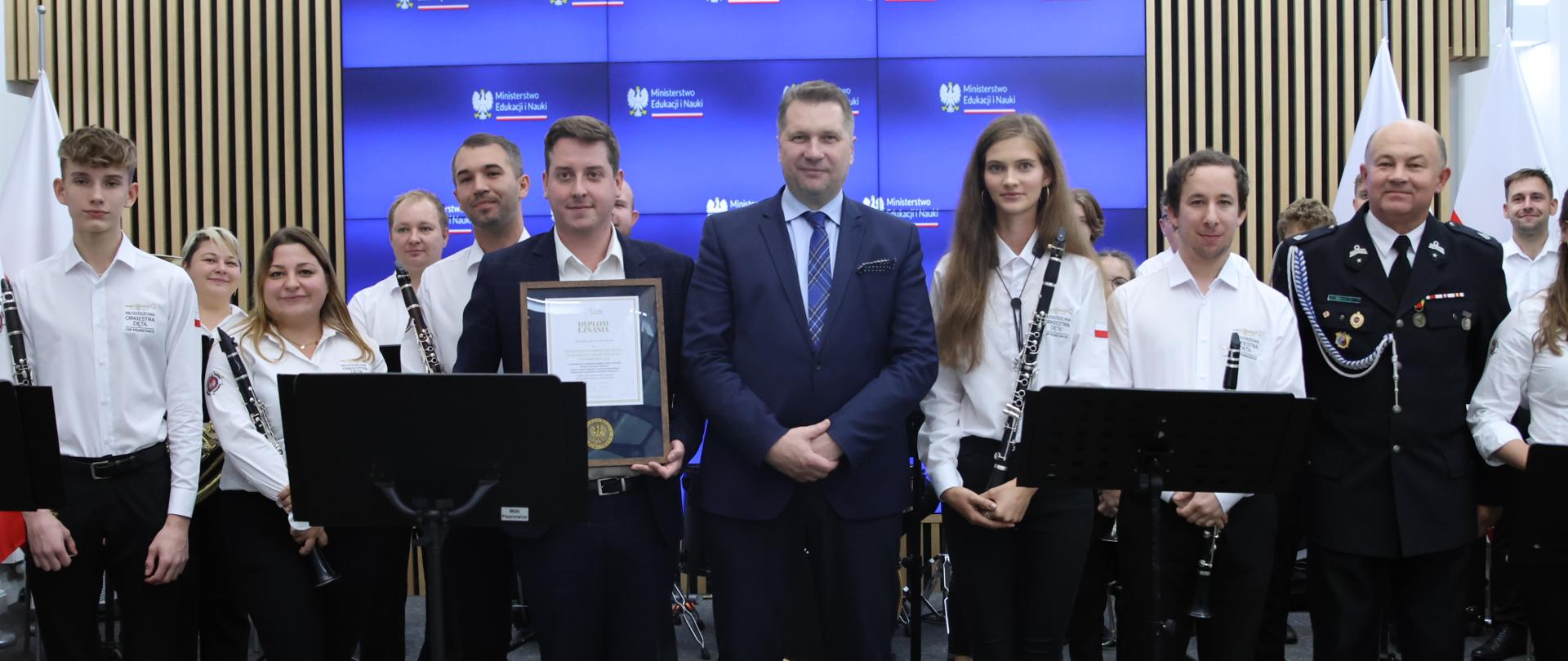W sali stoi minister Czarnek wśród kilkunastu młodych osób w białych koszulach, trzymających instrumenty muzyczne.