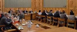 Widok z boku na dużą salę z zielonymi ścianami wyłożonymi częściowo drewnem, przy ustawionych w czworokąt drewnianych stołach siedzi dużo ludzi.