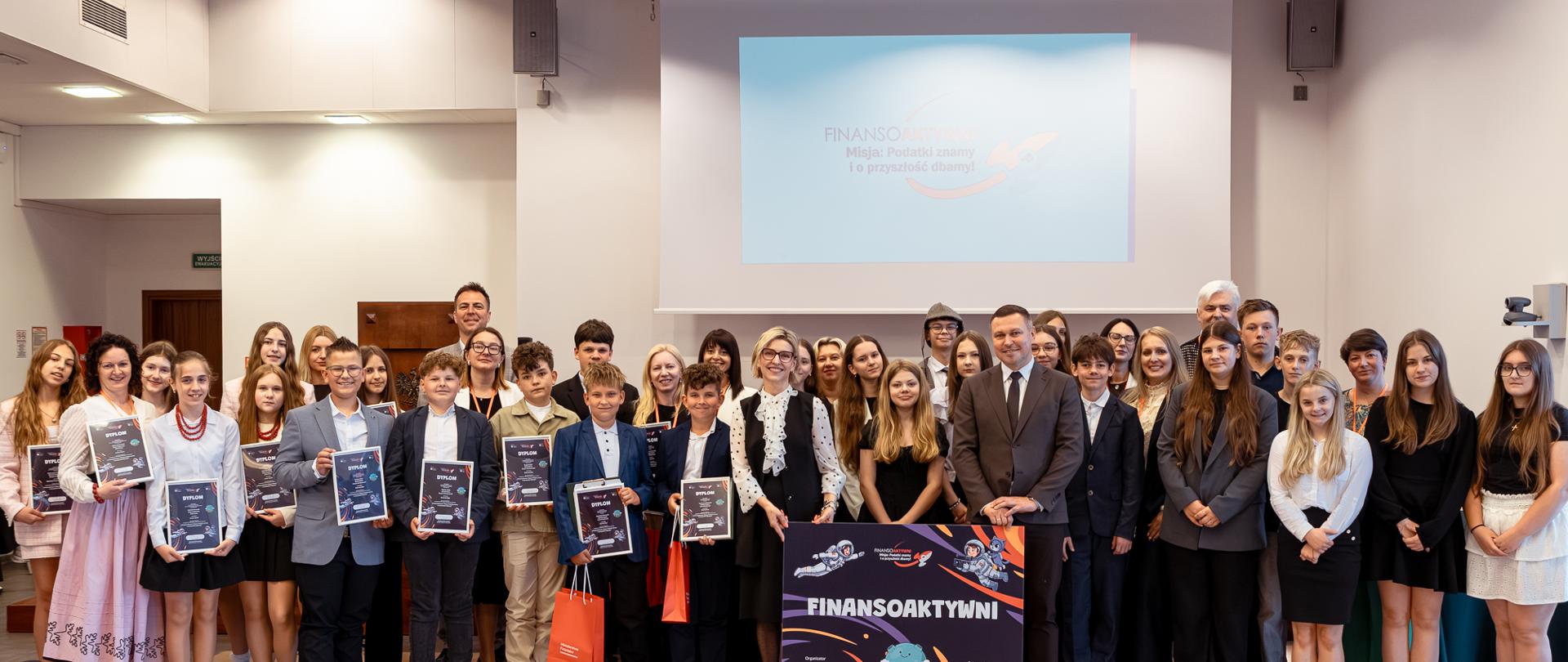 Grupa osób stoi w sali konferencyjnej, część trzyma dyplomy, a na środku widoczny jest plakat „FINANSOAKTYWNI” przed ekranem projekcyjnym.