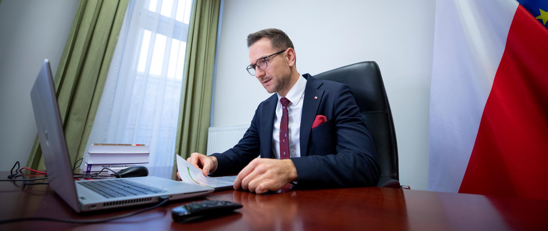 Wiceminister Waldemar Buda siedzi przy biurku przed laptopem. W ręku trzyma kartki. Za nim flaga Polski i UE.