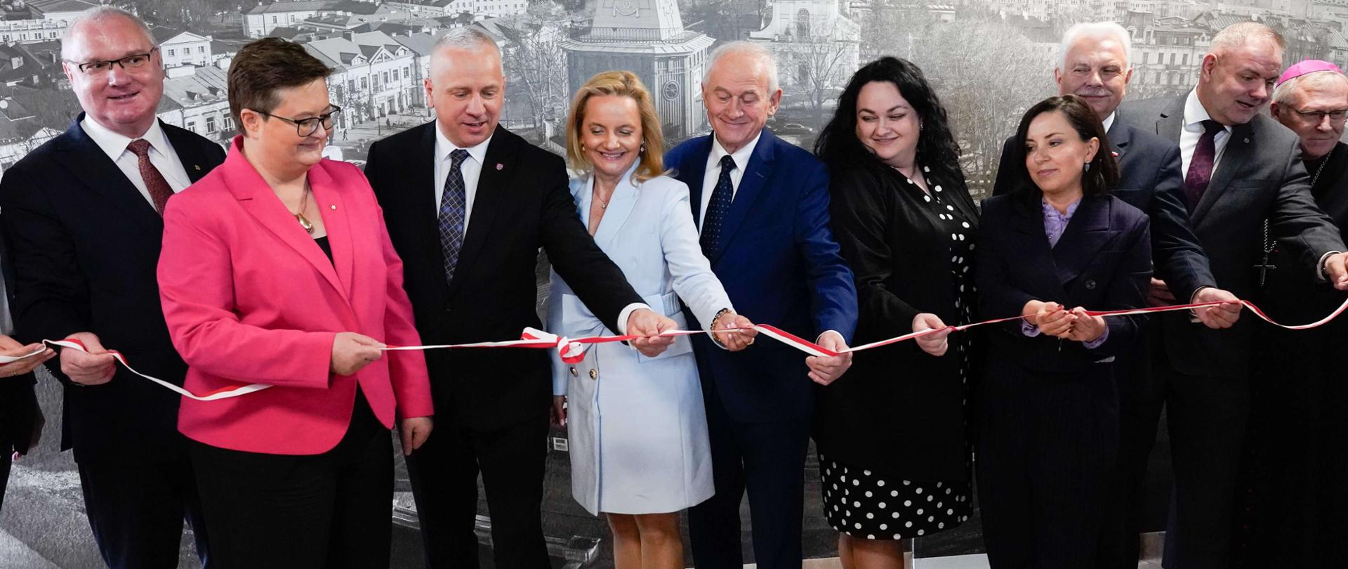 Dziewięć osób dorosłych stoi w rzędzie przed dużą, czarno-białą panoramą miasta, przecinając biało-czerwoną wstęgę. W środku grupy, kobieta w jasnoniebieskim płaszczu i mężczyzna w niebieskim garniturze trzymają nożyczki, przecinając wstęgę. Po ich lewej stronie stoi mężczyzna w okularach i różowej marynarce oraz mężczyzna w ciemnym garniturze. Po prawej stronie stoi kobieta w czarnej sukience w białe kropki oraz kobieta w ciemnym garniturze. Na obu końcach rzędu stoją mężczyźni w garniturach, a po prawej stronie widać także mężczyznę w sutannie i piusce.
Wszyscy uśmiechają się i patrzą na przecinaną wstęgę. W tle widoczna jest panorama miasta z charakterystyczną wieżą i innymi budynkami. Oświetlenie jest jasne, padające z góry. Atmosfera sugeruje uroczyste otwarcie lub inaugurację.