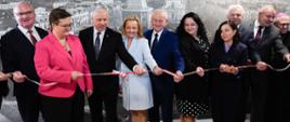 Dziewięć osób dorosłych stoi w rzędzie przed dużą, czarno-białą panoramą miasta, przecinając biało-czerwoną wstęgę. W środku grupy, kobieta w jasnoniebieskim płaszczu i mężczyzna w niebieskim garniturze trzymają nożyczki, przecinając wstęgę. Po ich lewej stronie stoi mężczyzna w okularach i różowej marynarce oraz mężczyzna w ciemnym garniturze. Po prawej stronie stoi kobieta w czarnej sukience w białe kropki oraz kobieta w ciemnym garniturze. Na obu końcach rzędu stoją mężczyźni w garniturach, a po prawej stronie widać także mężczyznę w sutannie i piusce.
Wszyscy uśmiechają się i patrzą na przecinaną wstęgę. W tle widoczna jest panorama miasta z charakterystyczną wieżą i innymi budynkami. Oświetlenie jest jasne, padające z góry. Atmosfera sugeruje uroczyste otwarcie lub inaugurację.