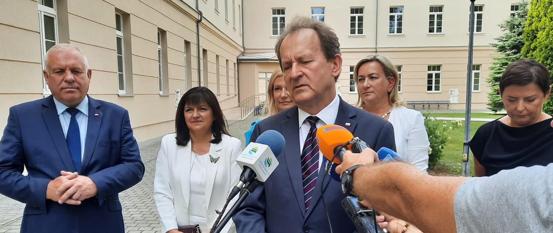 Na dworze przed budynkiem stoi grupa ludzi, z przodu wiceminister Bernacki mówi do kilku mikrofonów trzymanych przez reporterów.