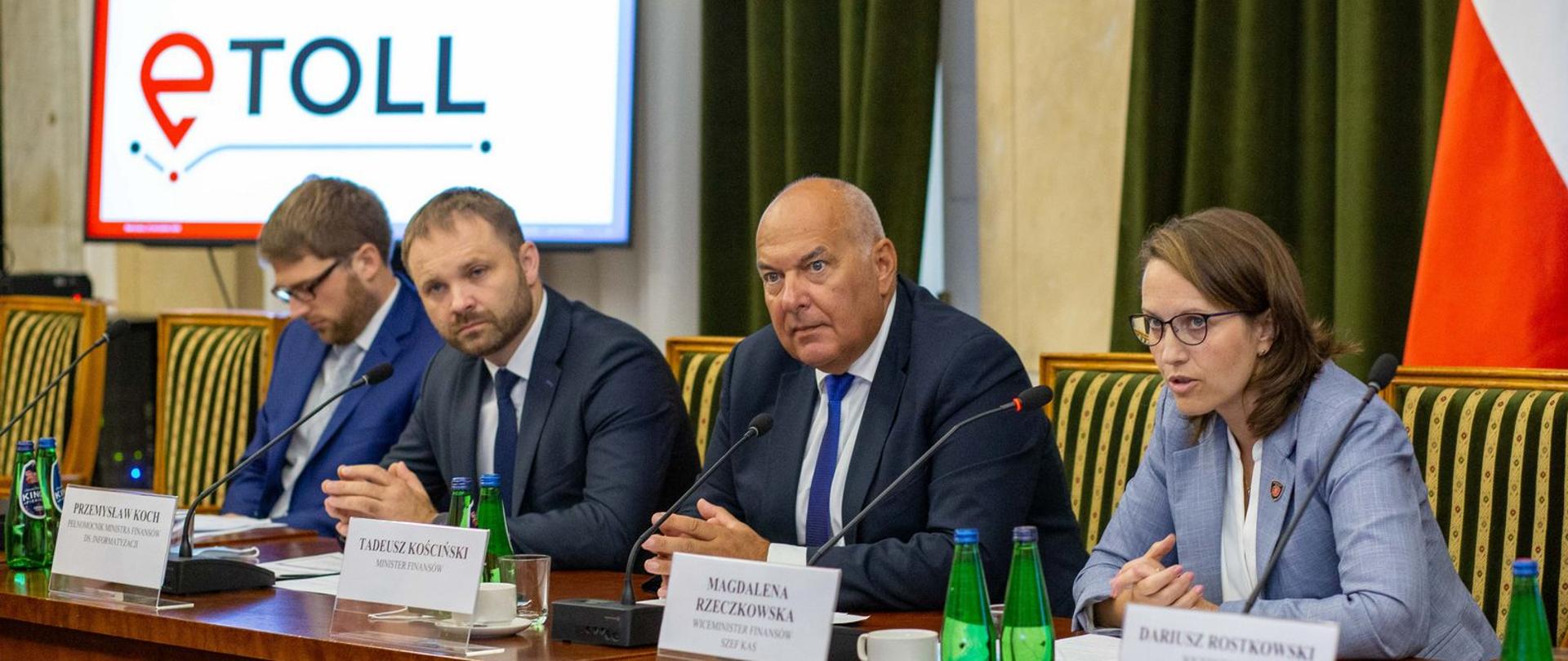 Stół prezydialny przy którym siedzą minister finansów Tadeusz Kościński, szefowa KAS Magdalena Rzeczkowska, pełnomocnik ministra finansów ds. informatyzacji Przemysław Koch, w tle slajd prezentacji z napisem e-Toll.