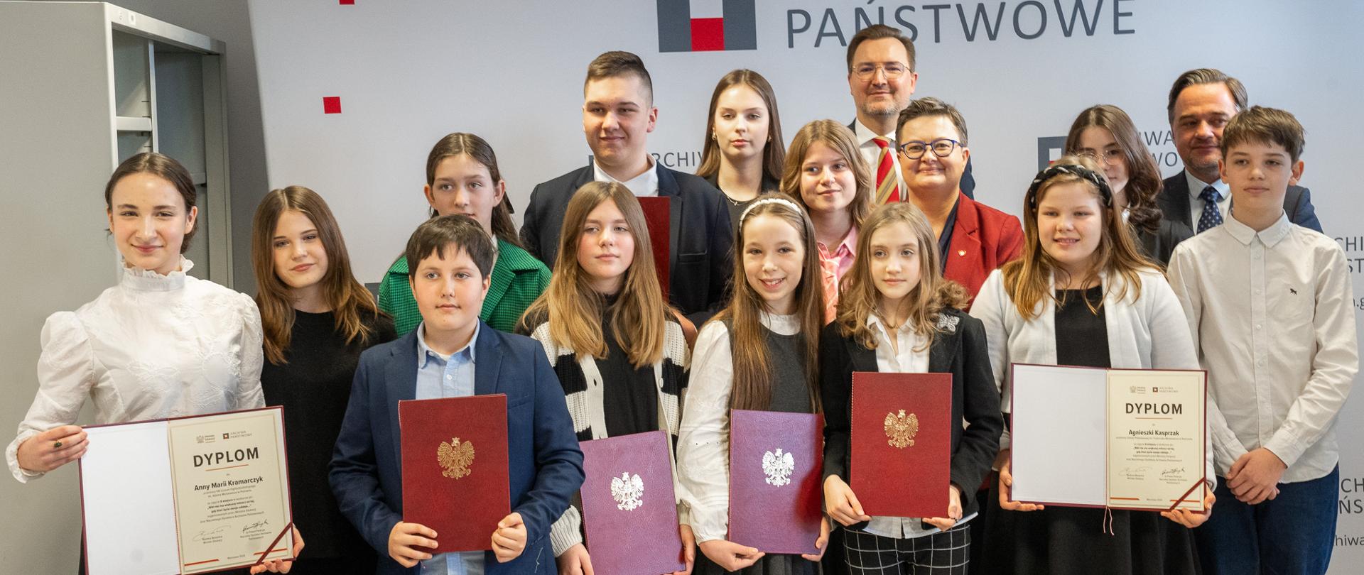Wspólne zdjęcie wiceminister Katarzyny Lubnauer, Naczelnego Dyrektora Archiwów Państwowych Paweła Pietrzyka oraz wszystkich laureatów i wyróżnionych uczestników konkursu. Uczestnicy stojący w pierwszym rzędzie pośrodku prezentują dyplomy zamknięte w czerwonej teczce ze złotym symbolem orzełka, natomiast dwie uczestniczki stojące po skrajnej lewej i prawej stronie trzymają teczki otwarte, pokazując dyplom. 