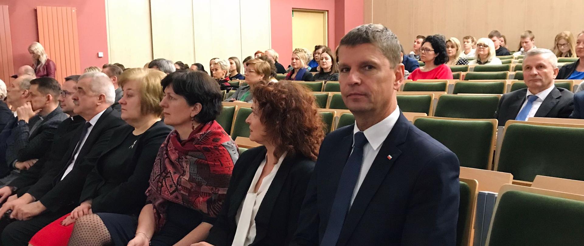 Widok z boku na dużą salę, na obitych na zielono rzędach krzeseł siedzą ludzie, w pierwszym rzędzie wiceminister Piontkowski, za nim kilka kobiet.