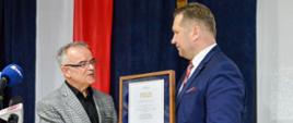 Minister Czarnek podaje mężczyźnie w marynarce w szarą kratę dokument oprawiony w ramki, za nimi ściana przysłonięta granatowym materiałem na środku którego wisi polska flaga.