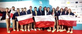 Na tle niebieskiej ściany stoi grupa kilkunastu osób w granatowych marynarkach, trzymają przed sobą trzy rozpostarte polskie flagi.