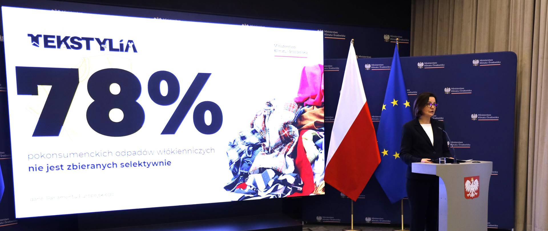 Wiceministra Anita Sowińska na konferencji w sali ministerstwa o nowych zasadach selektywnej zbiórki tekstyliów. Wiceministra stoi przy mównicy na tle polskich i europejskich flag i na tle telebimu. Na telebimie prezentacja pod tytułem "78 % pokonsumenckich odpadów włókienniczych nie jest zbieranych selektywnie".
