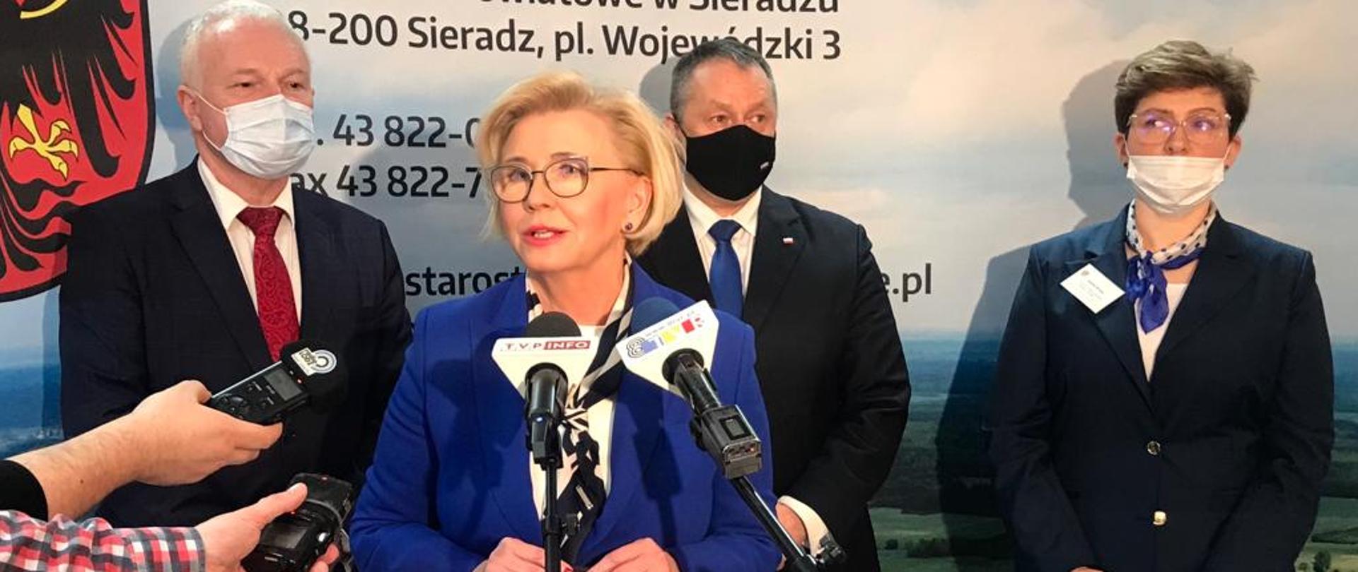 Wiceminister Marzena Machałek mówi podczas konferencji prasowej, stoi przy mikrofonach, za nią stoją inni uczestnicy konferencji.