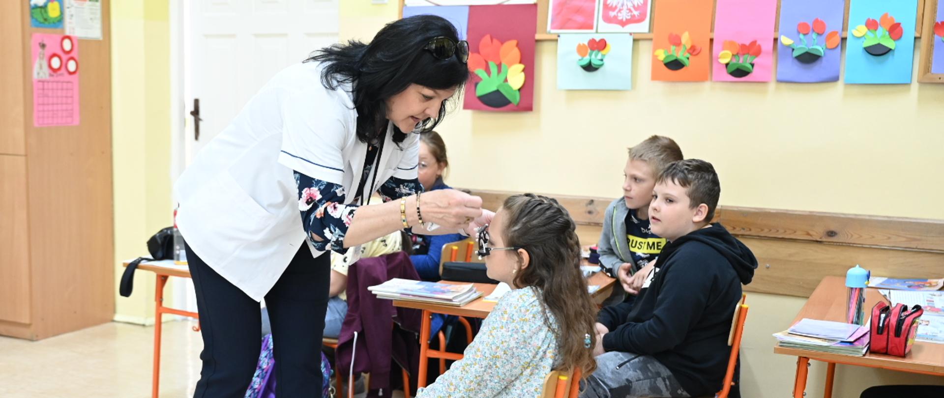 Uczennica jest przygotowywana do badania. Oprócz niej w sali lekcyjnej obecna jest lekarka oraz inne dzieci.