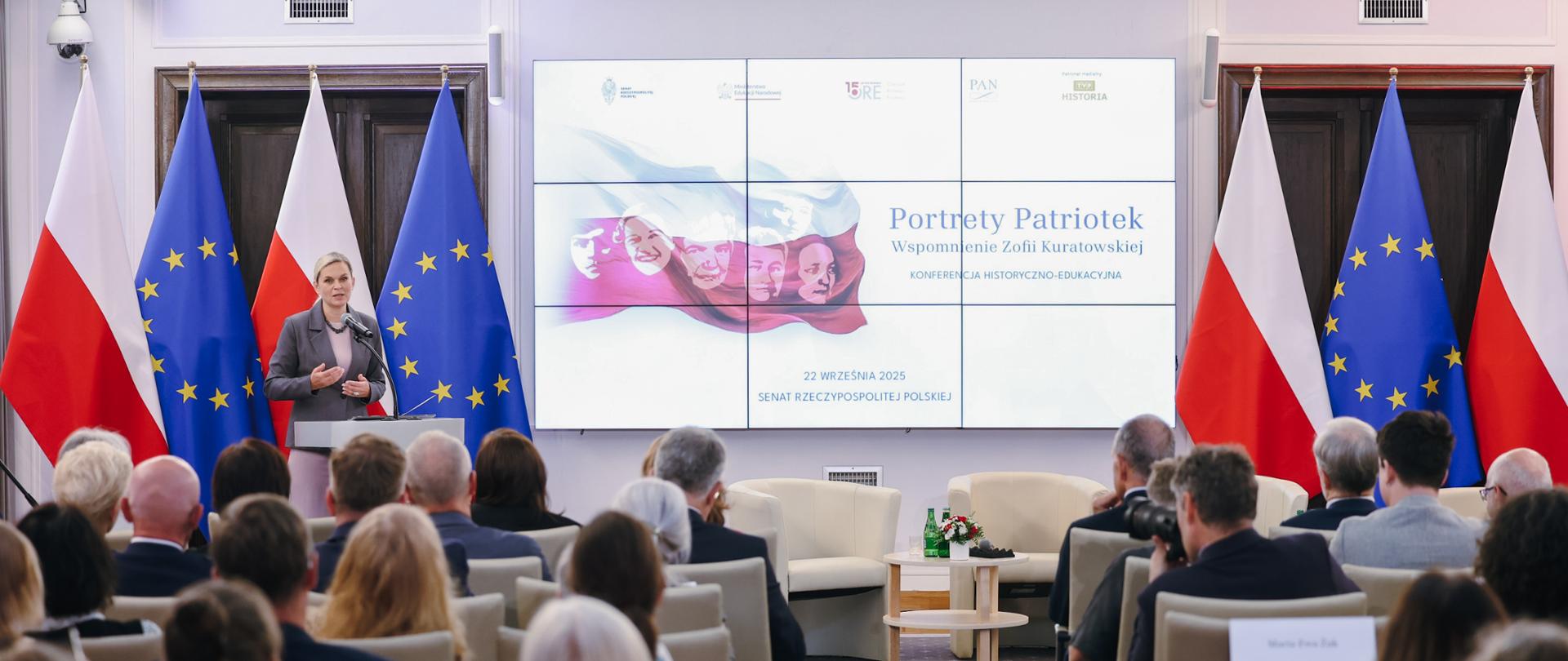 Kobieta z krótkimi, blond włosami, ubrana w szary garnitur, stoi za mównicą i przemawia do dużej grupy osób siedzących na krzesłach. Za nią znajdują się flagi Polski i Unii Europejskiej, a także duży ekran, na którym wyświetlany jest tytuł „Portrety Patriotek. Wspomnienie Zofii Kuratowskiej”. Na ekranie widoczne są również zdjęcia twarzy pięciu kobiet. Publiczność, składająca się z wielu osób, siedzi zwrócona w stronę mówczyni. W tle widać kamery monitoringu oraz inne detale wnętrza.