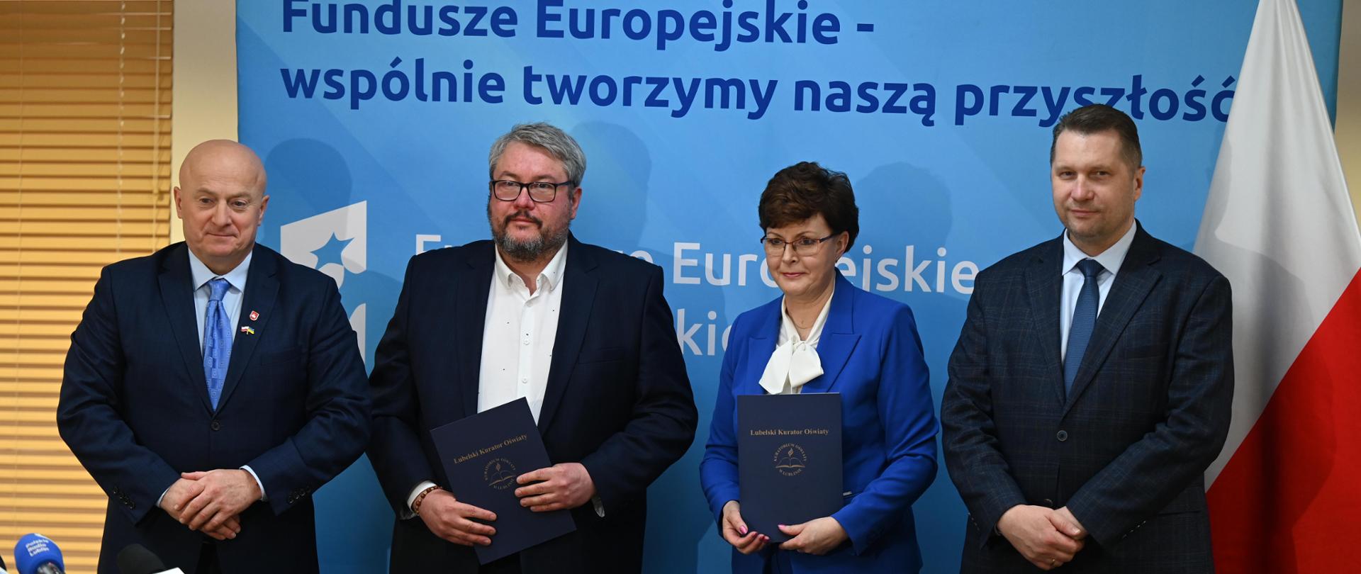 Za żółtym stołem stoją: minister Czarnek, kobieta w niebieskiej marynarce i dwóch mężczyzn w garniturach, na stole przed nimi stoi kilka mikrofonów, za nimi niebieska ściana z napisem Fundusz Europejskie - wspólnie tworzymy naszą przyszłość, z boku flaga Polski.