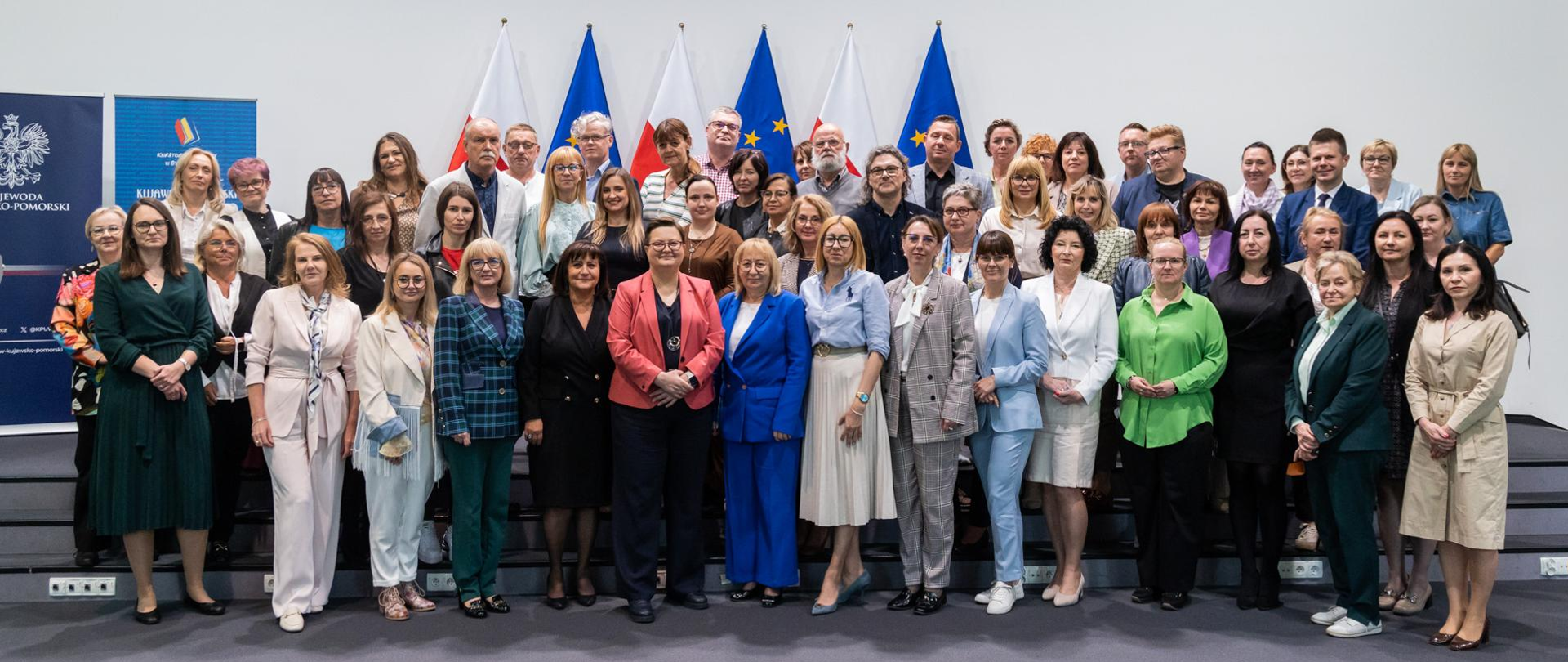 Na jasnym tle, przed rzędem flag Polski i Unii Europejskiej, stoi liczna grupa dorosłych, prawdopodobnie ponad 40 osób, pozujących do wspólnego zdjęcia. W pierwszym rzędzie stoją głównie kobiety, ubrane w różnorodne, eleganckie stroje, w tym sukienki, spódnice, bluzki i żakiety w wielu kolorach. Niektóre z nich trzymają w rękach dyplomy lub certyfikaty.
W drugim rzędzie, za pierwszym, stoją zarówno kobiety, jak i mężczyźni, również ubrani w formalne lub półformalne stroje. Wszyscy patrzą w stronę obiektywu i uśmiechają się. Po lewej stronie widoczny jest baner z polskim godłem i tekstem. Oświetlenie jest jasne, studyjne. Atmosfera sugeruje uroczyste wydarzenie, takie jak wręczenie nagród, konferencja lub pamiątkowe zdjęcie grupy współpracowników lub uczestników.