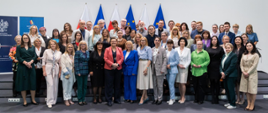 Na jasnym tle, przed rzędem flag Polski i Unii Europejskiej, stoi liczna grupa dorosłych, prawdopodobnie ponad 40 osób, pozujących do wspólnego zdjęcia. W pierwszym rzędzie stoją głównie kobiety, ubrane w różnorodne, eleganckie stroje, w tym sukienki, spódnice, bluzki i żakiety w wielu kolorach. Niektóre z nich trzymają w rękach dyplomy lub certyfikaty.
W drugim rzędzie, za pierwszym, stoją zarówno kobiety, jak i mężczyźni, również ubrani w formalne lub półformalne stroje. Wszyscy patrzą w stronę obiektywu i uśmiechają się. Po lewej stronie widoczny jest baner z polskim godłem i tekstem. Oświetlenie jest jasne, studyjne. Atmosfera sugeruje uroczyste wydarzenie, takie jak wręczenie nagród, konferencja lub pamiątkowe zdjęcie grupy współpracowników lub uczestników.
