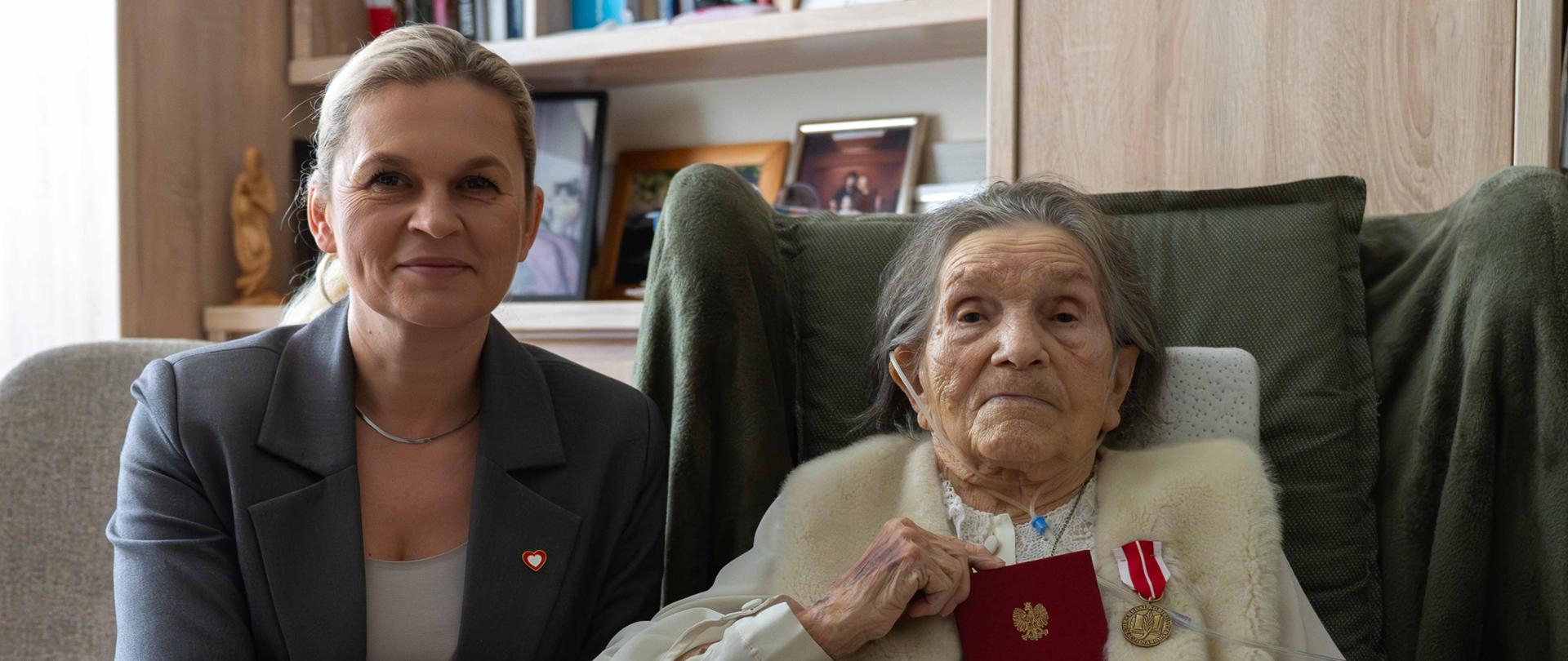 Barbara Nowacka siedzi przy fotelu Wandy Traczyk-Stawskiej. Obie kobiety patrzą w stronę aparatu. W tle widać domowe wnętrze z szafą z książkami i pamiątkami.