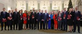 W dużej sali stoi duża grupa elegancko ubranych osób, za nimi kolumny, w górze wielki kryształowy żyrandol.