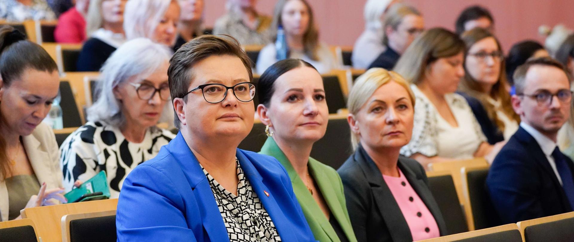Obraz przedstawia dużą grupę osób siedzących w rzędach na widowni lub w auli. Na pierwszym planie widać kilka kobiet i jednego mężczyznę, którzy patrzą w stronę, z której robiono zdjęcie. Kobieta w centrum na pierwszym planie ma na sobie niebieską marynarkę i okulary. W tle widać więcej osób siedzących na krzesłach.