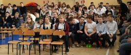 Widok od przodu na salę gimnastyczną, na krzesłach i ławkach ustawionych w rzędy siedzi wiele ubranych na galowo dzieci, pod ścianami drabinki, z tyłu sali kosz do koszykówki i bramka do piłki nożnej.