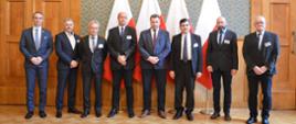 Ośmiu mężczyzn w garniturach, wśród nich minister Czarnek, stoi na tle zielonej ściany, za nimi cztery polskie flagi i drewniane drzwi.